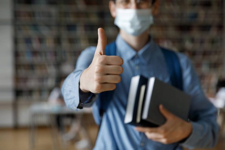 A student holding books and wearing a medical mask gives a thumbs visiting a college campus.