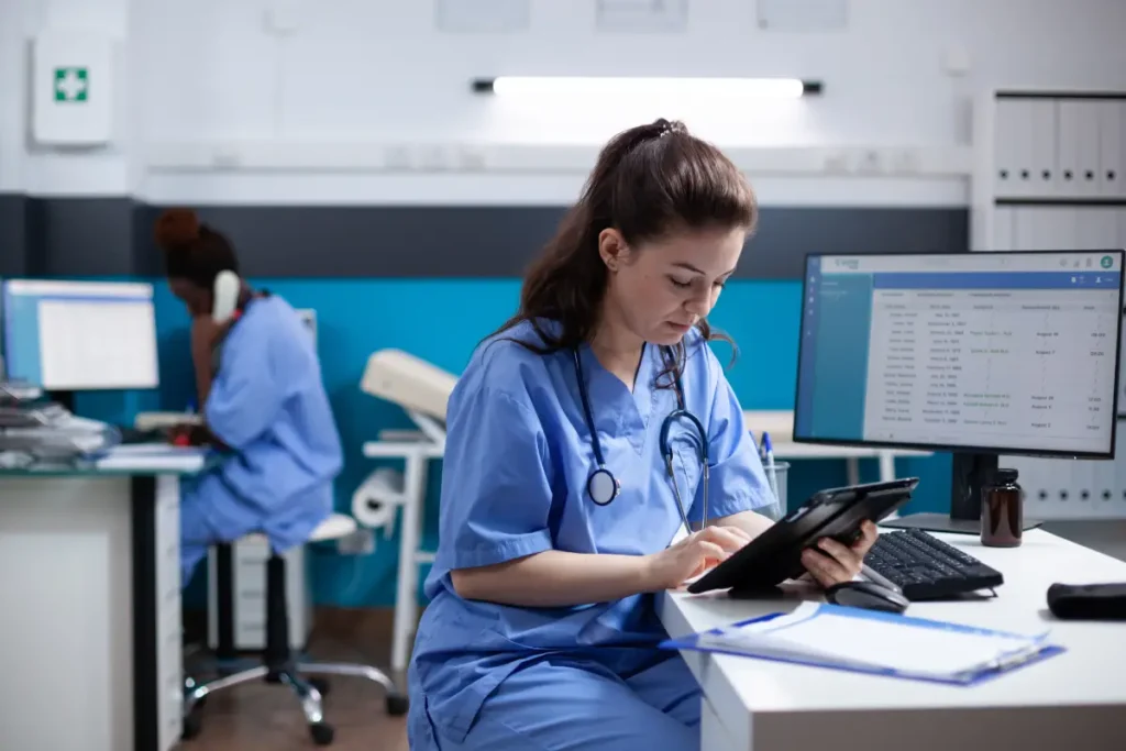 A practical nurse in scrubs reviews patient records on a tablet at her desk.