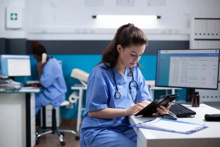 A practical nurse in scrubs reviews patient records on a tablet at her desk.